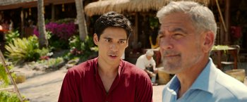 Movie still from “Ticket to Paradise” (2022), directed by Ol Parker – A young man in a red shirt is looking at the camera while an older man in a blue shirt looks on; Close Up shot, Over the shoulder angle