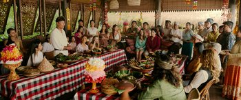Movie still from “Ticket to Paradise” (2022), directed by Ol Parker – A group of people sitting around a table with food on top of it; Wide shot, Over the shoulder angle