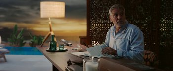 Movie still from “Ticket to Paradise” (2022), directed by Ol Parker – An older man sitting at a table writing on a piece of paper; Medium shot, Over the shoulder angle