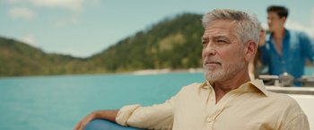 Movie still from “Ticket to Paradise” (2022), directed by Ol Parker – An older man sitting on a boat in front of a body of water; Close Up shot, Low angle