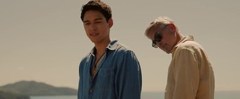 Movie still from “Ticket to Paradise” (2022), directed by Ol Parker – A man standing next to another man on a beach; Close Up shot, Over the shoulder angle