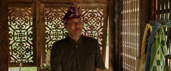 Movie still from “Ticket to Paradise” (2022), directed by Ol Parker – An older man wearing an oriental hat in front of a window; Medium shot, Over the shoulder angle