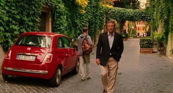 Movie still from “To Rome with Love” (2012), directed by Woody Allen – A man walking down the street in front of a red car; Wide shot, Over the shoulder angle