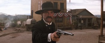 Movie still from “Tombstone” (1993), directed by George P. Cosmatos – A man in a black suit and a black hat is holding a gun; Medium shot, Low angle