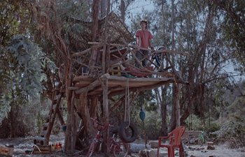 Movie still from “Too Late to Die Young” (2018), directed by Dominga Sotomayor – A man sitting on top of a wooden structure in the middle of a forest; Wide shot, Low angle