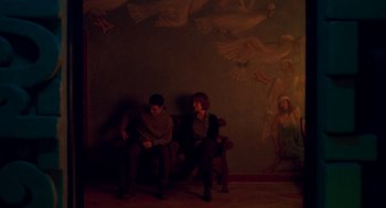 Movie still from “Too Old to Die Young” (2019), directed by Nicolas Winding Refn – Two people sitting on a chair in a room; Wide shot, Low angle