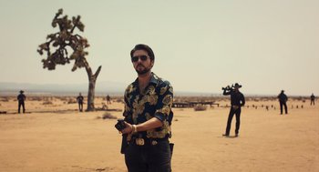 Movie still from “Too Old to Die Young” (2019), directed by Nicolas Winding Refn – A man standing in the desert holding a camera; Medium shot, Low angle