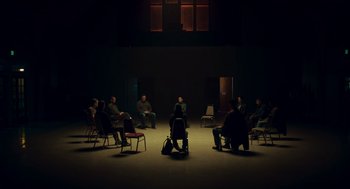 Movie still from “Too Old to Die Young” (2019), directed by Nicolas Winding Refn – A group of people sitting in chairs in a dark room; Extreme Wide shot, High angle
