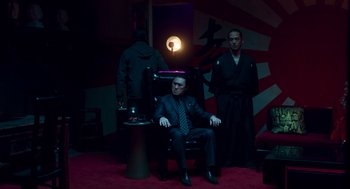 Movie still from “Too Old to Die Young” (2019), directed by Nicolas Winding Refn – A man sitting in a chair in front of two other men; Wide shot, High angle
