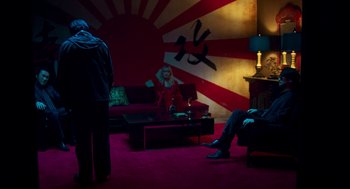 Movie still from “Too Old to Die Young” (2019), directed by Nicolas Winding Refn – People are sitting in a living room in front of a painting; Wide shot, Over the shoulder angle