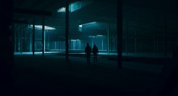 Movie still from “Too Old to Die Young” (2019), directed by Nicolas Winding Refn – Two people are standing in a dark building; Extreme Wide shot, Low angle