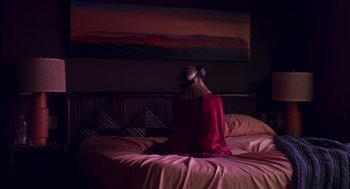 Movie still from “Too Old to Die Young” (2019), directed by Nicolas Winding Refn – A woman sitting on top of a bed in a room; Medium shot, Over the shoulder angle