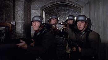 Movie still from “Top Secret!” (1984), directed by Jim Abrahams – A group of men wearing helmets and holding guns in front of a stone wall; Medium shot, Low angle