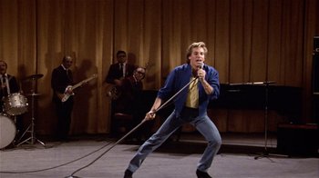 Movie still from “Top Secret!” (1984), directed by Jim Abrahams – A man holding a microphone while standing on a stage; Wide shot, Low angle