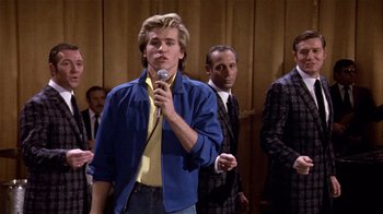 Movie still from “Top Secret!” (1984), directed by Jim Abrahams – A man holding a microphone while standing next to other men; Medium shot, Low angle