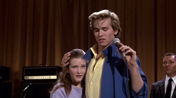 Movie still from “Top Secret!” (1984), directed by Jim Abrahams – A man and a woman standing next to each other holding microphones; Close Up shot, Over the shoulder angle