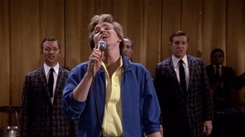 Movie still from “Top Secret!” (1984), directed by Jim Abrahams – A man singing into a microphone in front of a group of men; Medium shot, Low angle