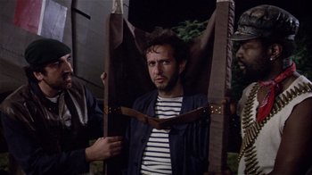 Movie still from “Top Secret!” (1984), directed by Jim Abrahams – A man in a striped shirt is being held up by two other men; Medium shot, Over the shoulder angle