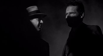 Movie still from “Touch of Evil” (1958), directed by Orson Welles – A man in a hat and a man in a suit standing next to each other in the dark; Close Up shot, Low angle