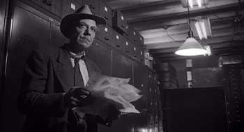 Movie still from “Touch of Evil” (1958), directed by Orson Welles – A man in a suit and hat holding papers in a room; Medium shot, Low angle