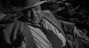 Movie still from “Touch of Evil” (1958), directed by Orson Welles – An old photo of a man holding a gun and wearing a hat; Close Up shot, Low angle