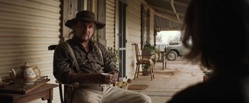 Movie still from “Tracks” (2013), directed by John Curran – An older man sitting on a porch holding a cup of coffee; Medium shot, Over the shoulder angle