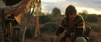 Movie still from “Tracks” (2013), directed by John Curran – A man sitting on the side of a road next to a bird cage; Medium shot, Over the shoulder angle