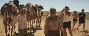 Movie still from “Tracks” (2013), directed by John Curran – A man walking down a dirt road next to a herd of camels; Wide shot, Over the shoulder angle