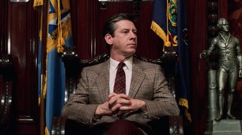 Movie still from “Trading Places” (1983), directed by John Landis – A man in a suit and tie sitting in a chair in front of flags; Medium shot, Low angle