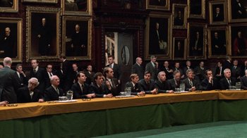 Movie still from “Trading Places” (1983), directed by John Landis – A group of men sitting at a long table in front of paintings; Wide shot, High angle