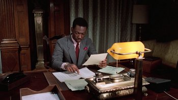 Movie still from “Trading Places” (1983), directed by John Landis – A man sitting at a table with papers and a lamp; Medium shot, High angle