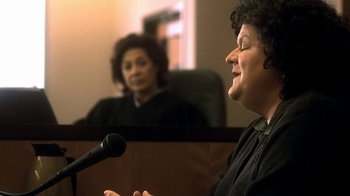 Movie still from “Traffic” (2000), directed by Steven Soderbergh – A woman in a courtroom with a microphone and another woman in the background; Close Up shot, Over the shoulder angle