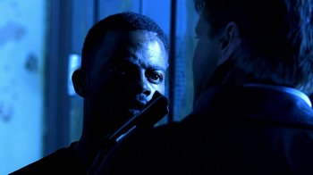Movie still from “Traffic” (2000), directed by Steven Soderbergh – A man holding a gun in front of another man in a dark room; Close Up shot, Over the shoulder angle