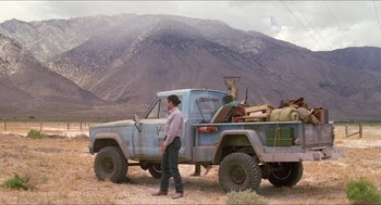 Movie still from “Tremors” (1990), directed by Ron Underwood – A man standing in front of a blue truck with mountains in the background; Wide shot, Low angle