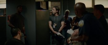 Movie still from “Triple 9” (2016), directed by John Hillcoat – A man in a black t - shirt is standing in front of some people; Medium shot, Over the shoulder angle