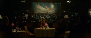 Movie still from “Triple 9” (2016), directed by John Hillcoat – A woman sitting at a table with a candle in front of a painting of an eagle; Wide shot, Low angle