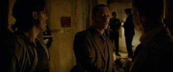 Movie still from “Triple 9” (2016), directed by John Hillcoat – A man wearing a suit and tie standing in a room; Medium shot, Over the shoulder angle