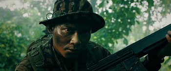 Movie still from “Triple Threat” (2019), directed by Jesse V. Johnson – A man in camouflage holding a rifle in a forest; Close Up shot, Low angle