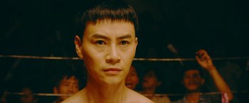 Movie still from “Triple Threat” (2019), directed by Jesse V. Johnson – A young man with a shaved head and no shirt in a boxing ring; Close Up shot, Over the shoulder angle