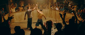 Movie still from “Triple Threat” (2019), directed by Jesse V. Johnson – Two men are fighting in a boxing ring while people watch; Wide shot, High angle