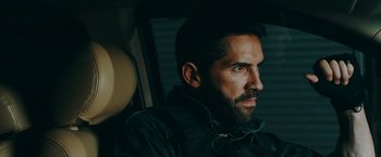 Movie still from “Triple Threat” (2019), directed by Jesse V. Johnson – A man sitting in the back seat of a car wearing ear buds; Close Up shot, Over the shoulder angle