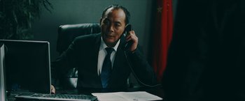 Movie still from “Triple Threat” (2019), directed by Jesse V. Johnson – A man sitting at a desk talking on a phone; Medium shot, Over the shoulder angle