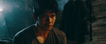 Movie still from “Triple Threat” (2019), directed by Jesse V. Johnson – A young man with dark curly hair wearing a brown shirt; Close Up shot, Over the shoulder angle