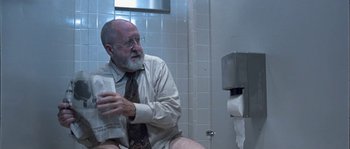 Movie still from “True Lies” (1994), directed by James Cameron – An older man sitting on a toilet in a bathroom; Medium shot, Low angle