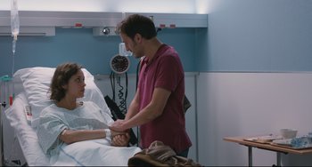 Movie still from “Two Days, One Night” (2014), directed by Jean-Pierre Dardenne – A man is holding a woman's arm in a hospital bed; Medium shot, Over the shoulder angle