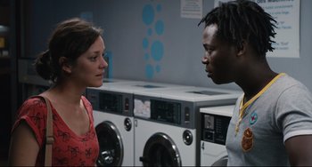 Movie still from “Two Days, One Night” (2014), directed by Jean-Pierre Dardenne – A man and a woman talking in front of each other in front of washing machines; Medium shot, Over the shoulder angle