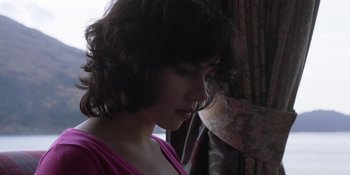 Movie still from “Under the Skin” (2013), directed by Jonathan Glazer – A young woman with dark curly hair wearing a pink shirt; Close Up shot, Low angle