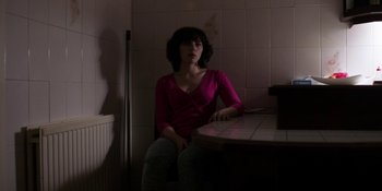 Movie still from “Under the Skin” (2013), directed by Jonathan Glazer – A woman sitting at a table in a dark room; Medium shot, Low angle