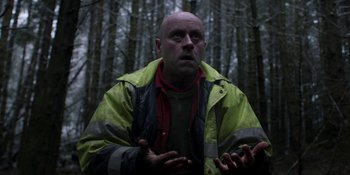 Movie still from “Under the Skin” (2013), directed by Jonathan Glazer – A bald man in a yellow and black jacket in the woods; Medium shot, Low angle