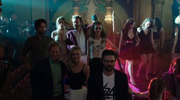 Movie still from “Urge” (2016), directed by Aaron Kaufman – A group of people posing for a picture in front of a stage; Wide shot, High angle